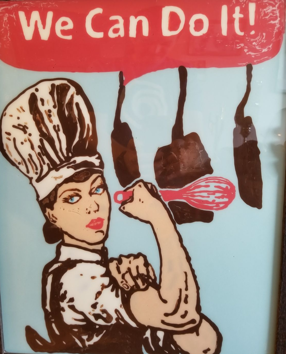A poster that depicts a female chef in a position of homage to Rosie the Riveter. The poster is made out of coloured chocolate: red, white, brown and blue.
