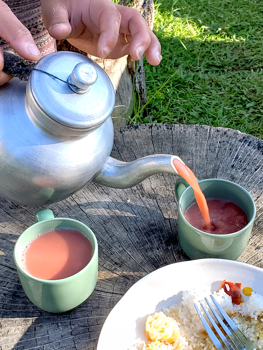 Close up of hands pouring yak butter tea from a teapot into two green cups sitting on a tree stump, witha plate of rice and other food adjacent.